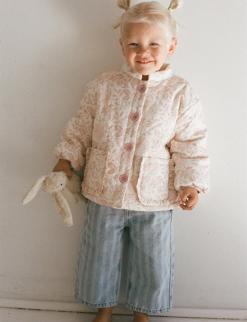 Child wearing a floral jacket and blue pants, holding a stuffed animal against a white wall.
