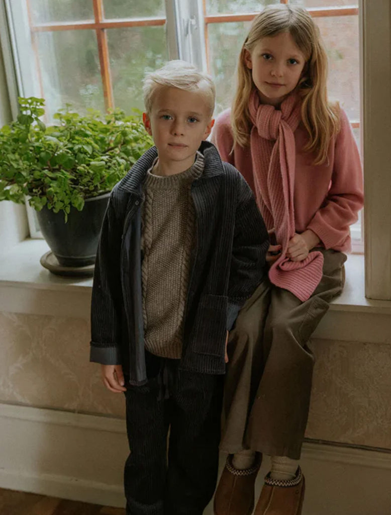 Two children standing indoors by a window with plants in the background