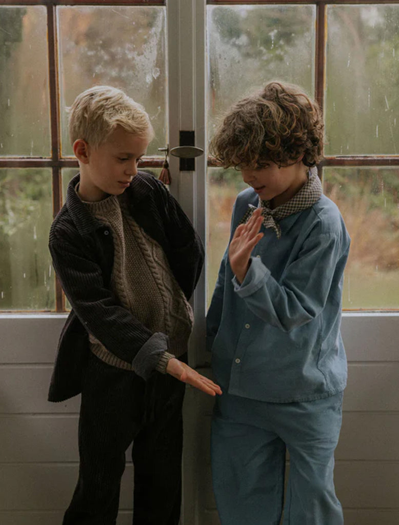 Two children standing indoors near a glass door with a blurred outdoor background