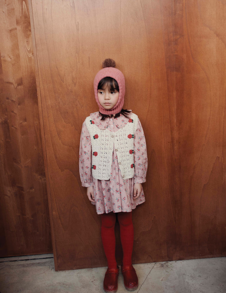 Child wearing a pink outfit with a white cardigan against a wooden wall