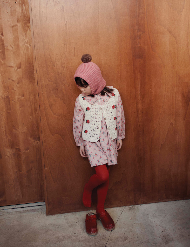 Child wearing a pink knitted hat, white lace cardigan, floral dress, red tights, and brown shoes against a wooden wall.