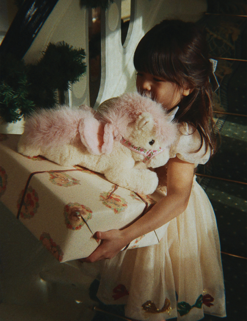 Young girl holding a teddy bear and a wrapped gift in a softly lit room.