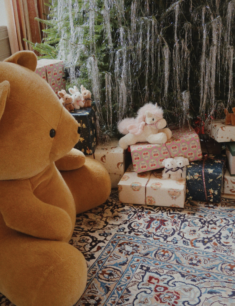 Large teddy bear sitting on a patterned rug with Christmas decorations in the background.