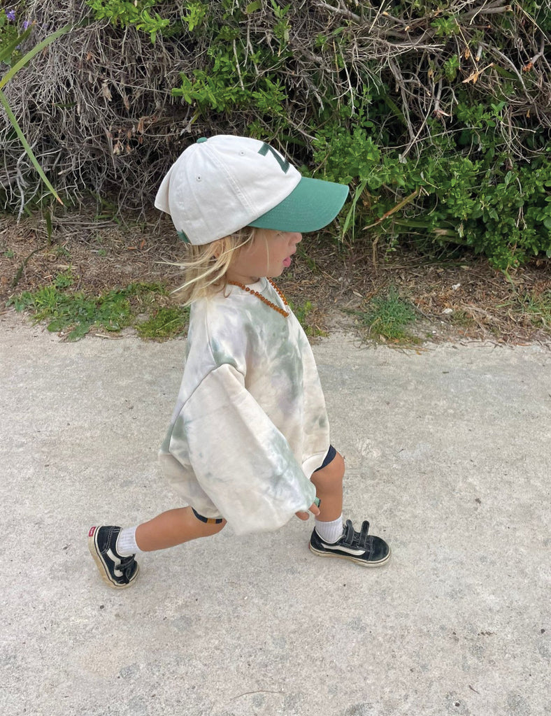 Child walking outdoors wearing a tie-dye shirt, white cap, and green visor.