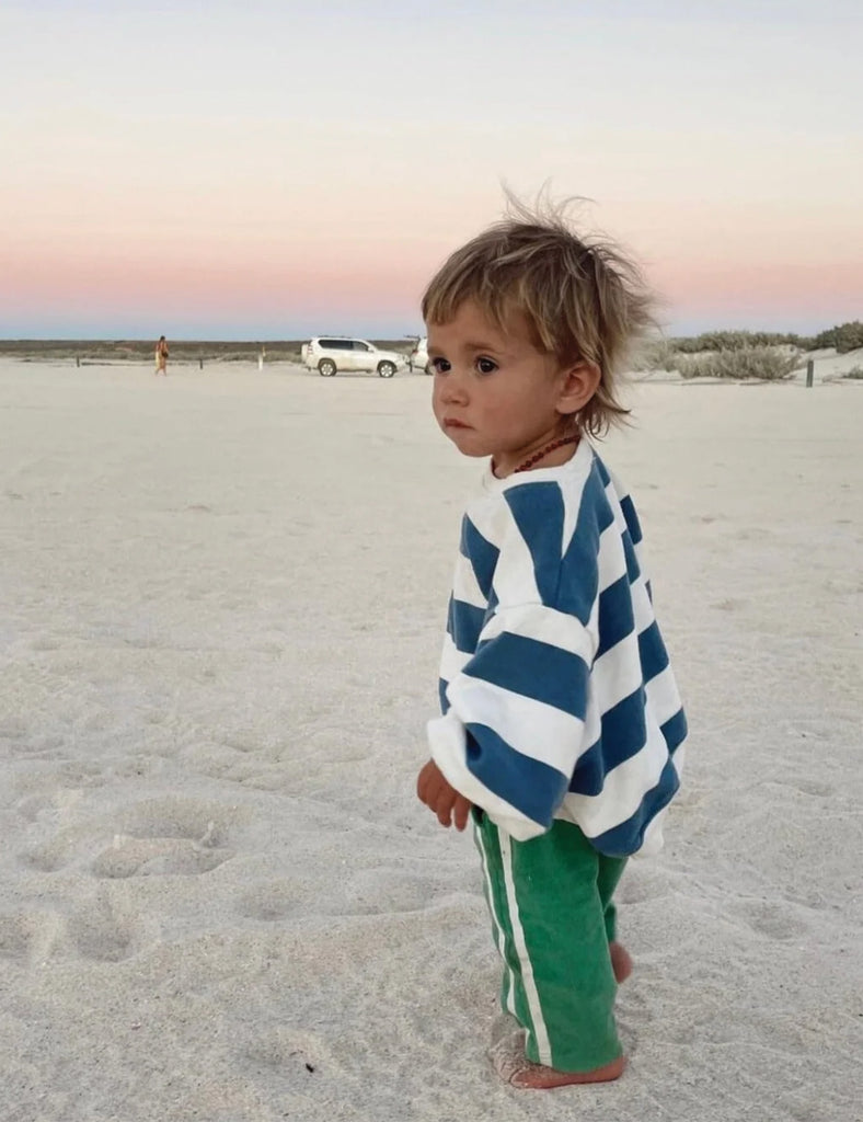 Child wearing a blue and white striped poncho on a sandy beach with a car and people in the distance.