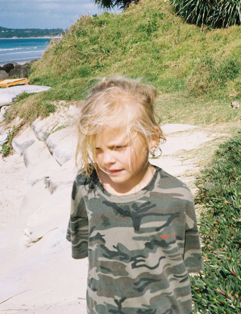Child in a camouflage shirt standing on a beach with grass and water in the background