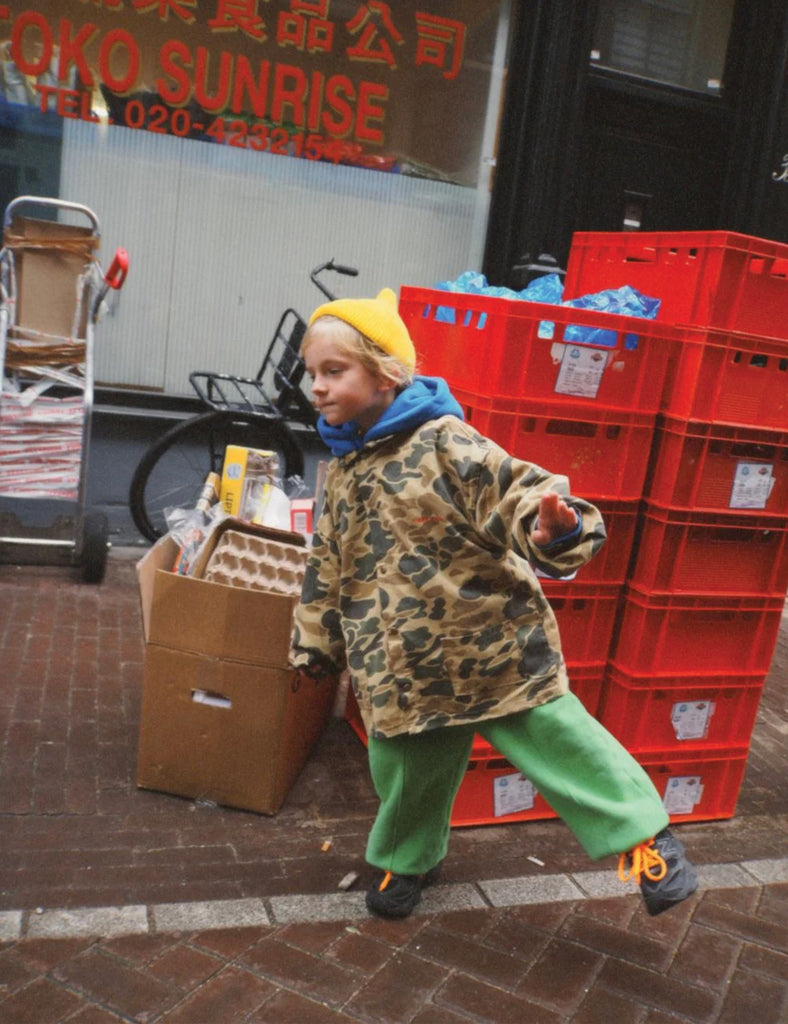 Child in a camouflage jacket and green pants standing next to red crates and a cardboard box on a street.