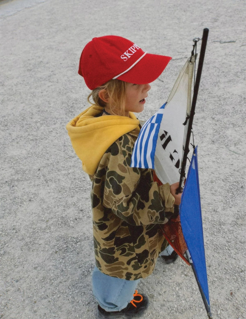 Child wearing a red cap and camouflage jacket holding a kite on a paved surface