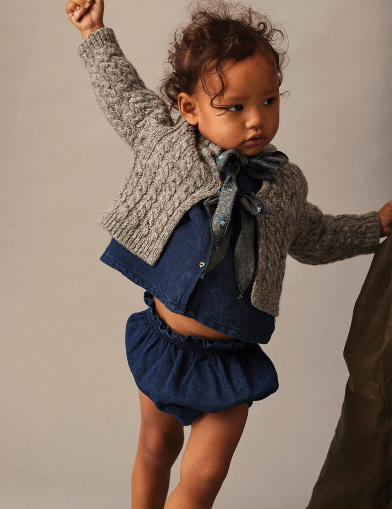 Child wearing a gray knitted cardigan over a blue outfit with a bow, on a plain background