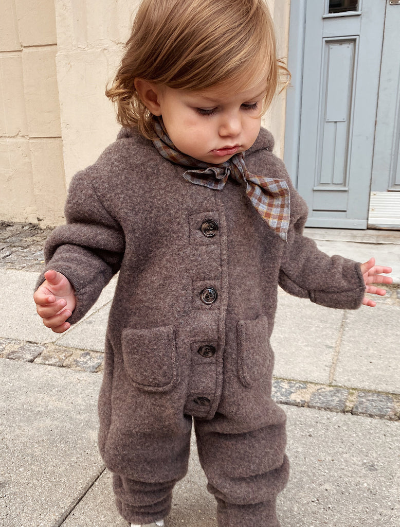Child wearing a brown coat with buttons and plaid scarf outdoors.