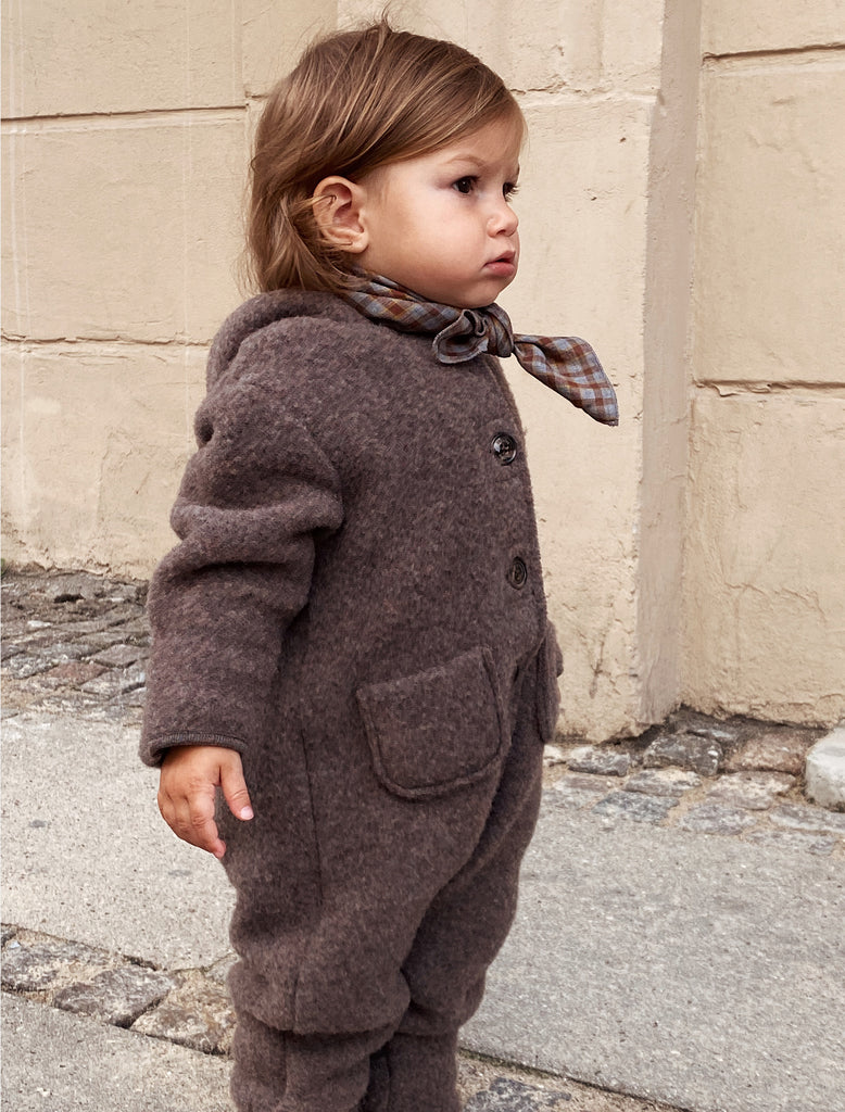 Child wearing a brown coat with a plaid scarf standing against a beige wall.
