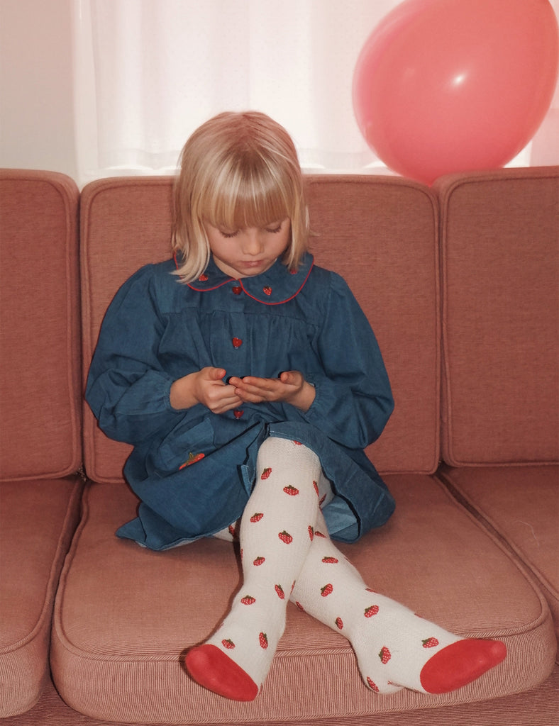 Child sitting on a couch wearing a blue dress and white tights with red patterns, surrounded by pink balloons.