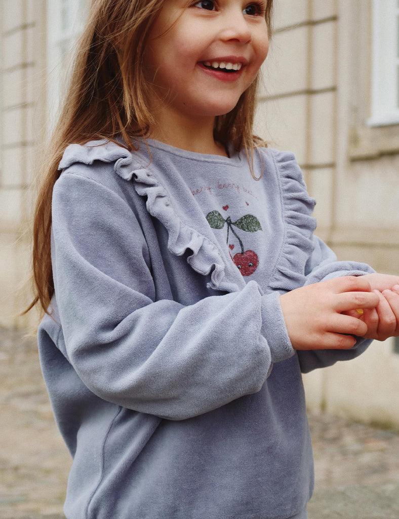 Young girl wearing a light blue sweater with ruffled sleeves and floral design, standing outdoors.